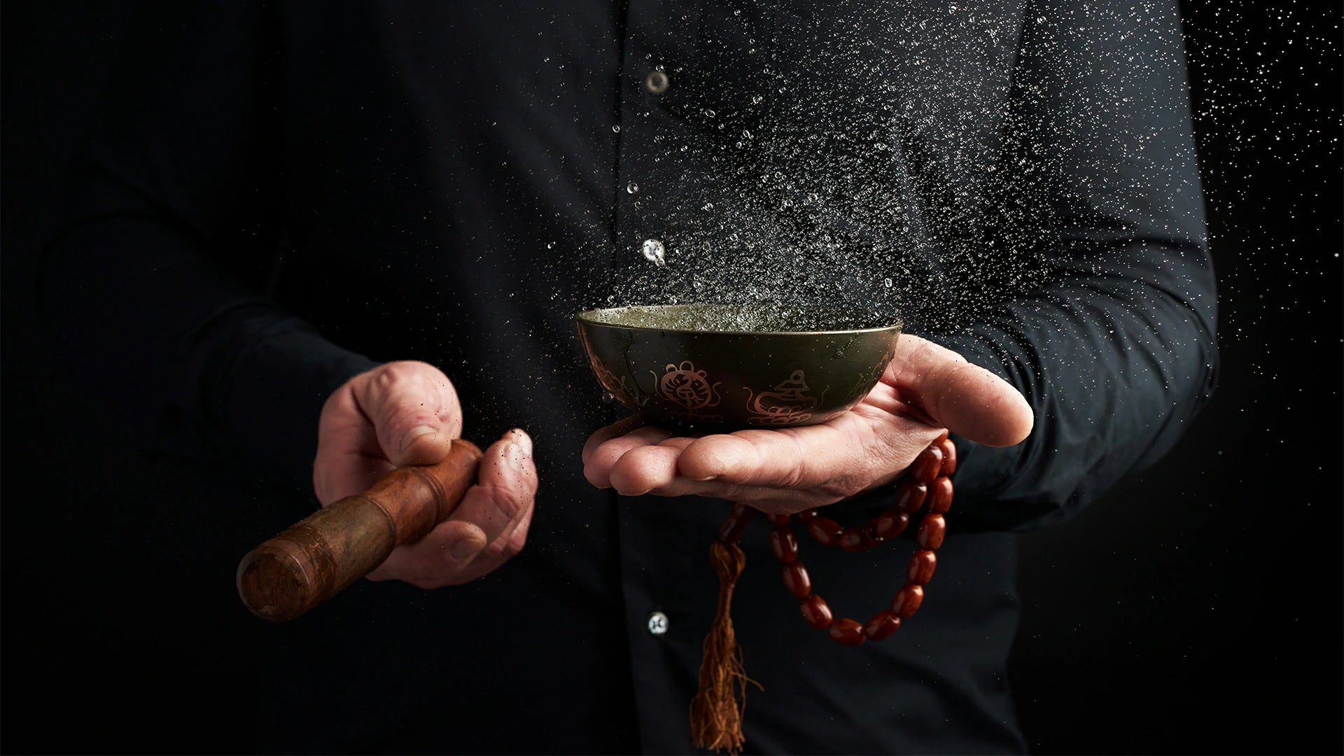 A man in a black shirt and bracelets hits a singing bowl, splashing water